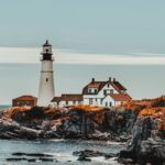 Lighthouse near big house near orange grass on rocky shore near ocean under bright sky in daytime
